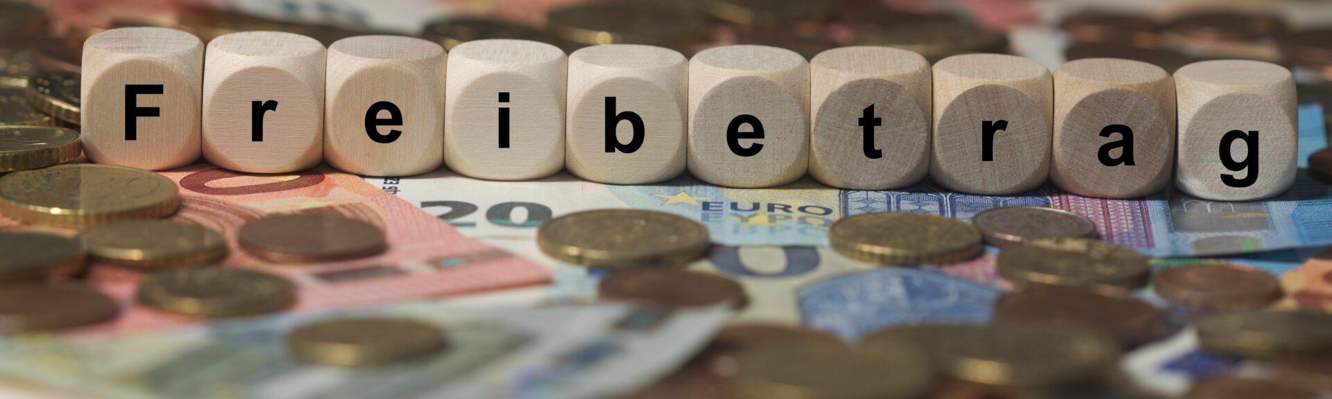 freibetrag - Holzwürfel mit Buchstaben im Hintergrund mit Geld, Geldscheine