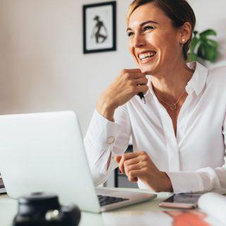 Woman sitting at her desk in office holding a pen. Smiling business woman with her laptop computer on her office desk.
