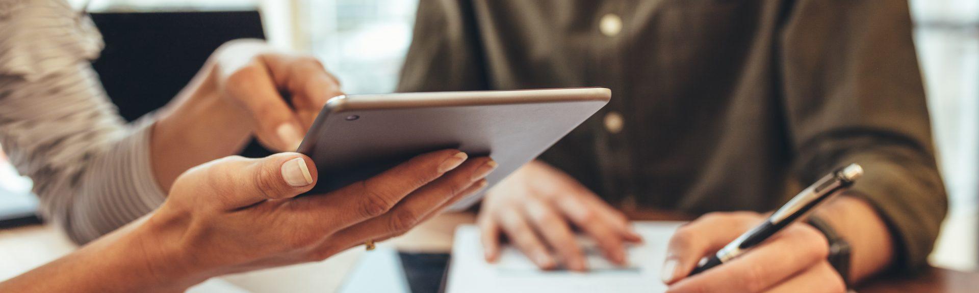 Woman showing a tablet computer to her colleague while discussing work in office. Man writing notes in book sitting at his work desk.