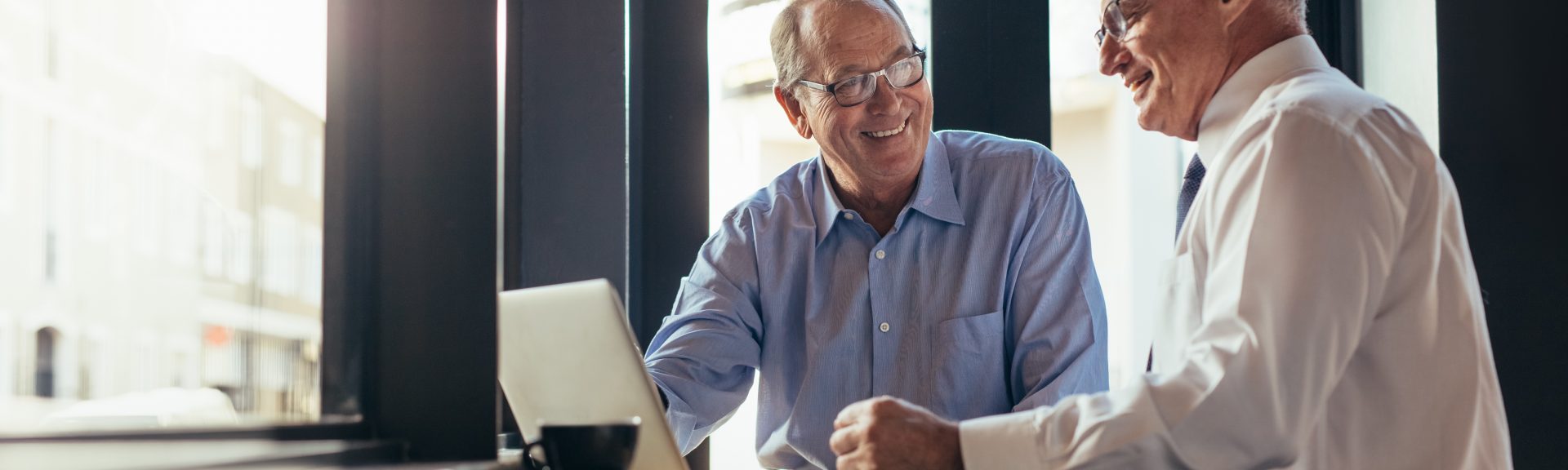 Two senior business men working together in modern cafe. Business partners sitting near window with laptop on table at restaurant.