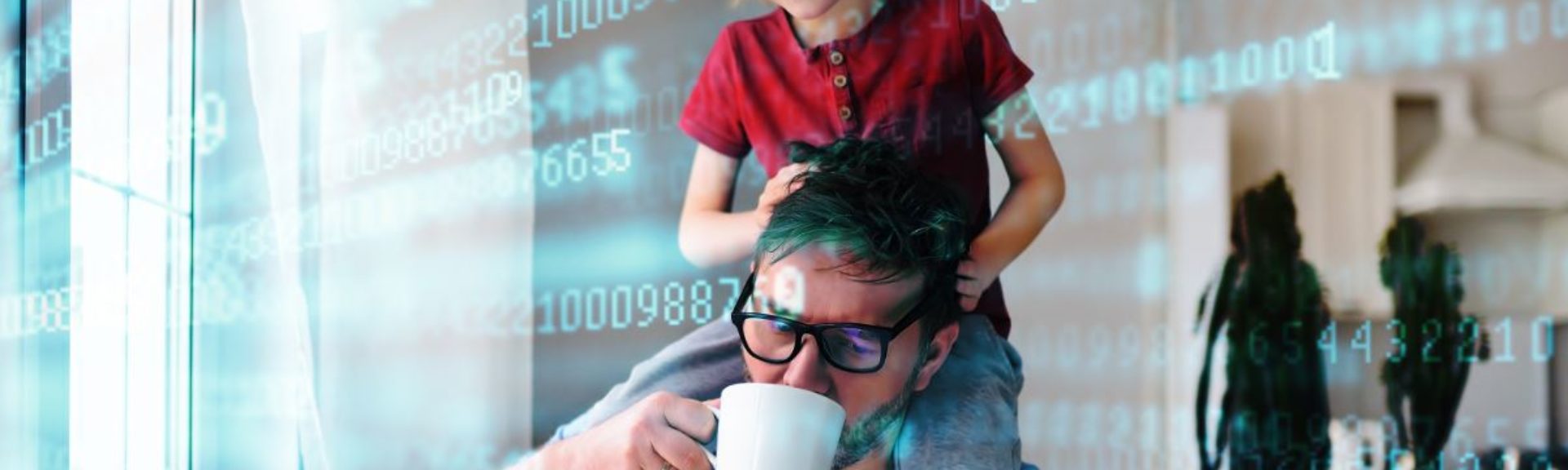 Dad in Homeoffice with a coffee mug in his hand sitting at the table with his laptop and his son is sitting on his shoulders