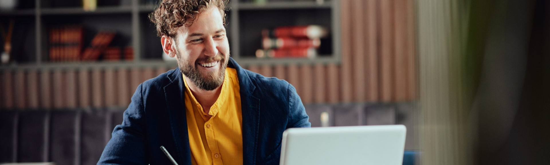 Young smiling bearded Caucasian blogger dressed smart casual writing notes in agenda and looking at laptop while sitting in cafeteria.