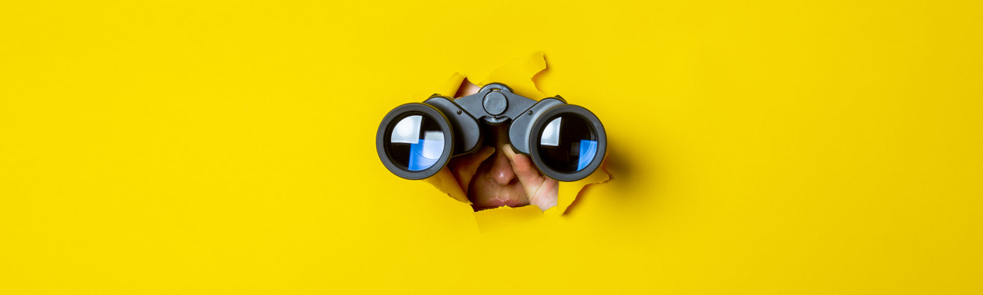 Female hand holds black binoculars on a yellow background. Journey, find and search concept.