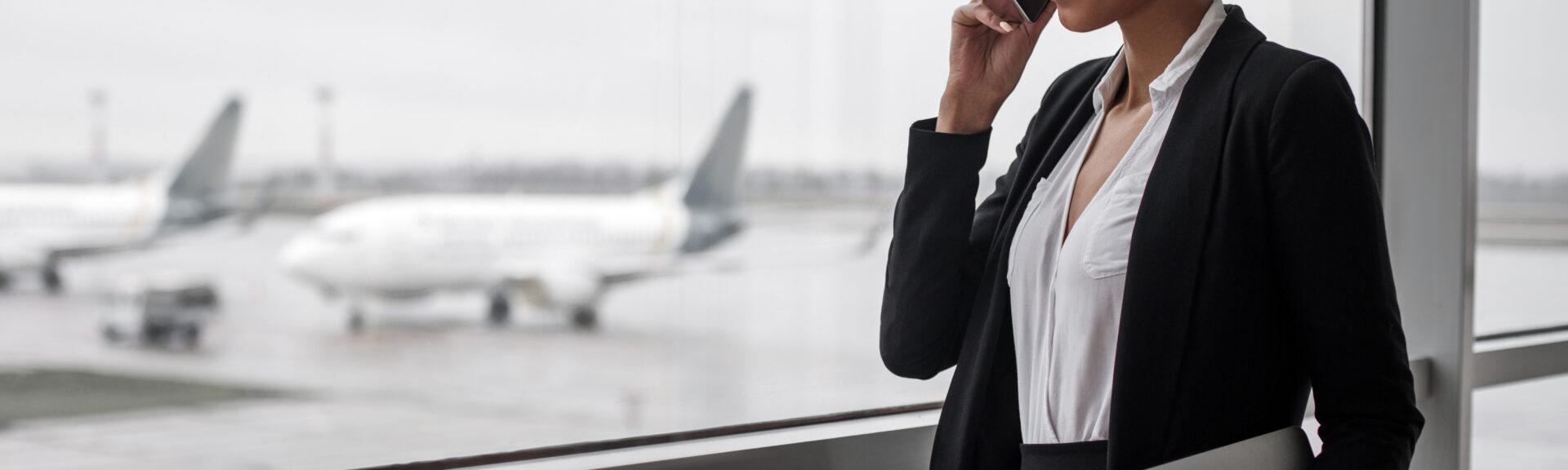 Work everywhere. Side view of young smiling woman is standing at airport lounge with laptop and looking through the window while enjoying conversation on mobile. Copy space and aircrafts in background