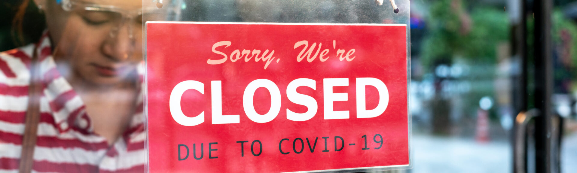 An upset Local small business owner woman wearing a protective face shield turning the sign for closed in front of the cafe, coffee shop, or store glass door cause lockdown quarantine due to covid-19.