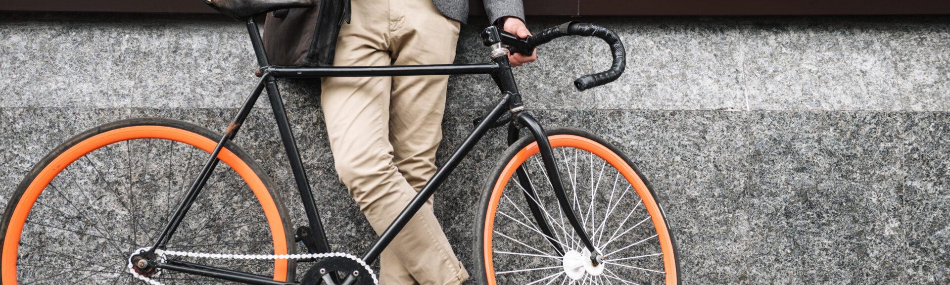 Cropped image of young businessman leaning on a wall while standing outdoors with a bicycle and using mobile phone