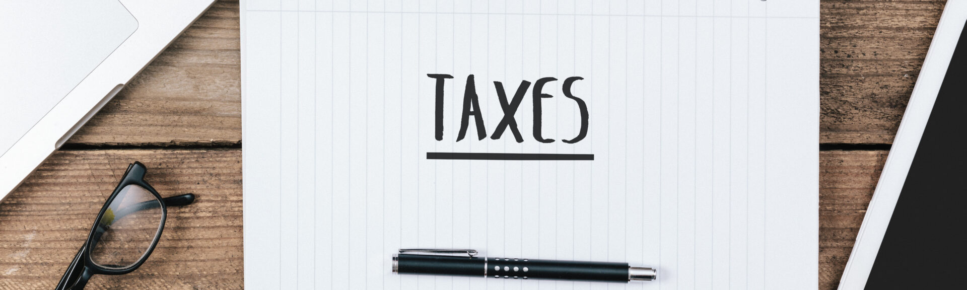 Taxes text on note padoffice desk with electronic devices, computer and paper, wood table from above, concept image for blog title or header image.