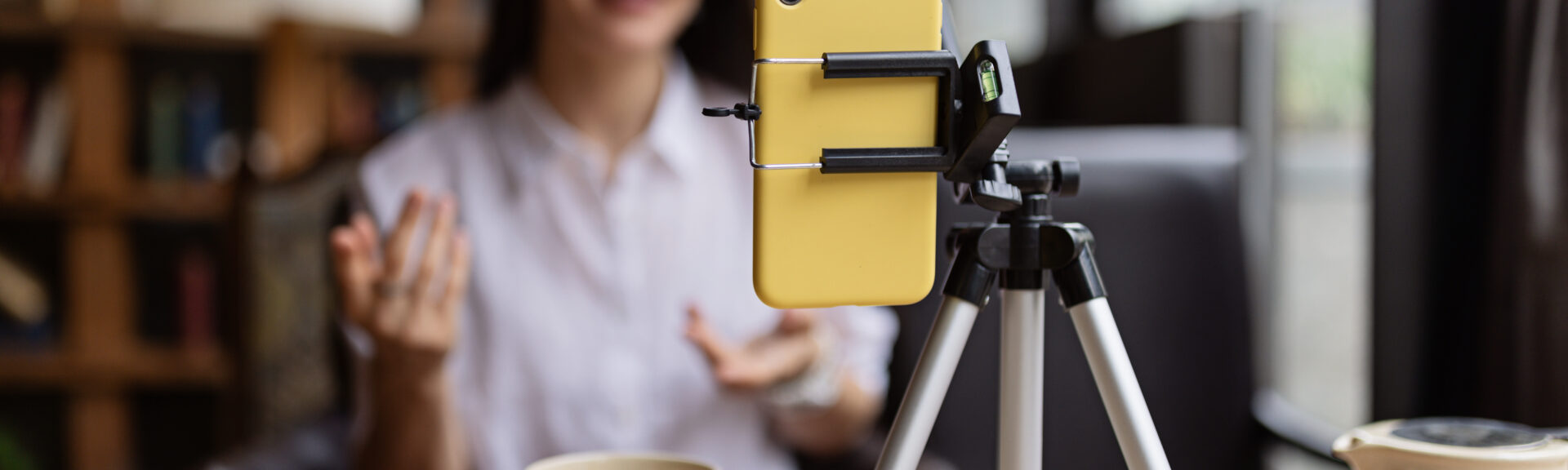 Happy young Caucasian millennial or gen z woman with long brunette hair streaming with smart phone on tripod, shooting social media blog in modern cafe. Influencer using social networks